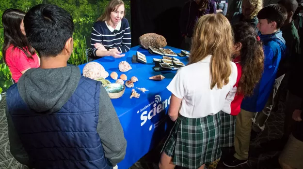 Boys and girls look at snail specimens on a table where zoologist Ellen Strong is sitting. She is speaking to the students.