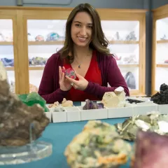 A woman holding a reddish-pink mineral while standing at a table that has many other minerals, and gems on it.