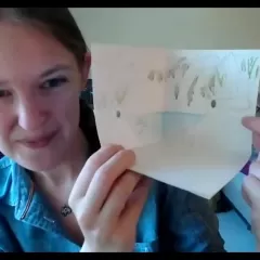 Ingrid Rochon, a light-skinned woman wearing a blue shirt, holds up a piece of paper with drawings on it