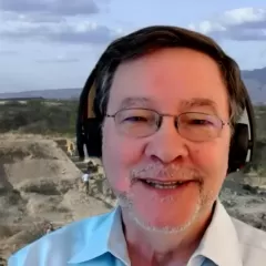 A man's face with an African landscape in the background. He has short brown hair, glasses, and a short gray beard and he is wearing headphones.