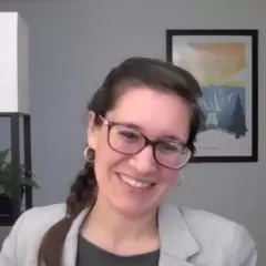 A smiling woman with braided dark hair. She is wearing glasses, a gray jacket, and a black shirt. Behind her is a framed painting on a wall.