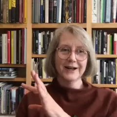 A blonde-haired woman wearing glasses and a brown shirt, with a bookcase behind her.