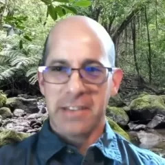 A balding, light-skinned man with glasses in front of a forest background