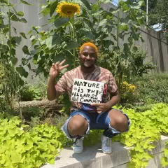 a black man with orange hair kneeling under giant sunflower with a sign reads nature is the pillar of community