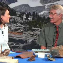 Maggy Benson and Dr. Bill Fitzhugh are seated at a table holding some rocks, antlers, tools, and a book.