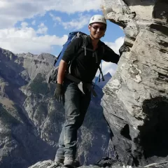 A man leaning against a rock face while wearing a helmet and backpack. Mountains are in the background.