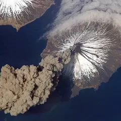 Aerial view of an ash plume rising from Mount Cleveland volcano, which has snow on its upper slopes.