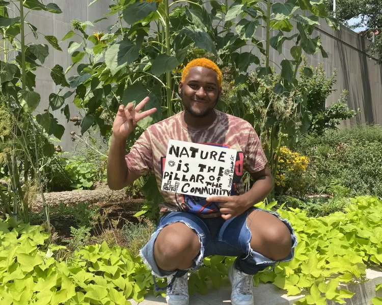 a black man with orange hair kneeling under giant sunflower with a sign reads nature is the pillar of community