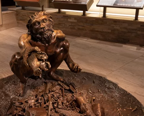 Bronze sculpture of an early human with a beard and mustache squatting and holding out a piece of wood. 