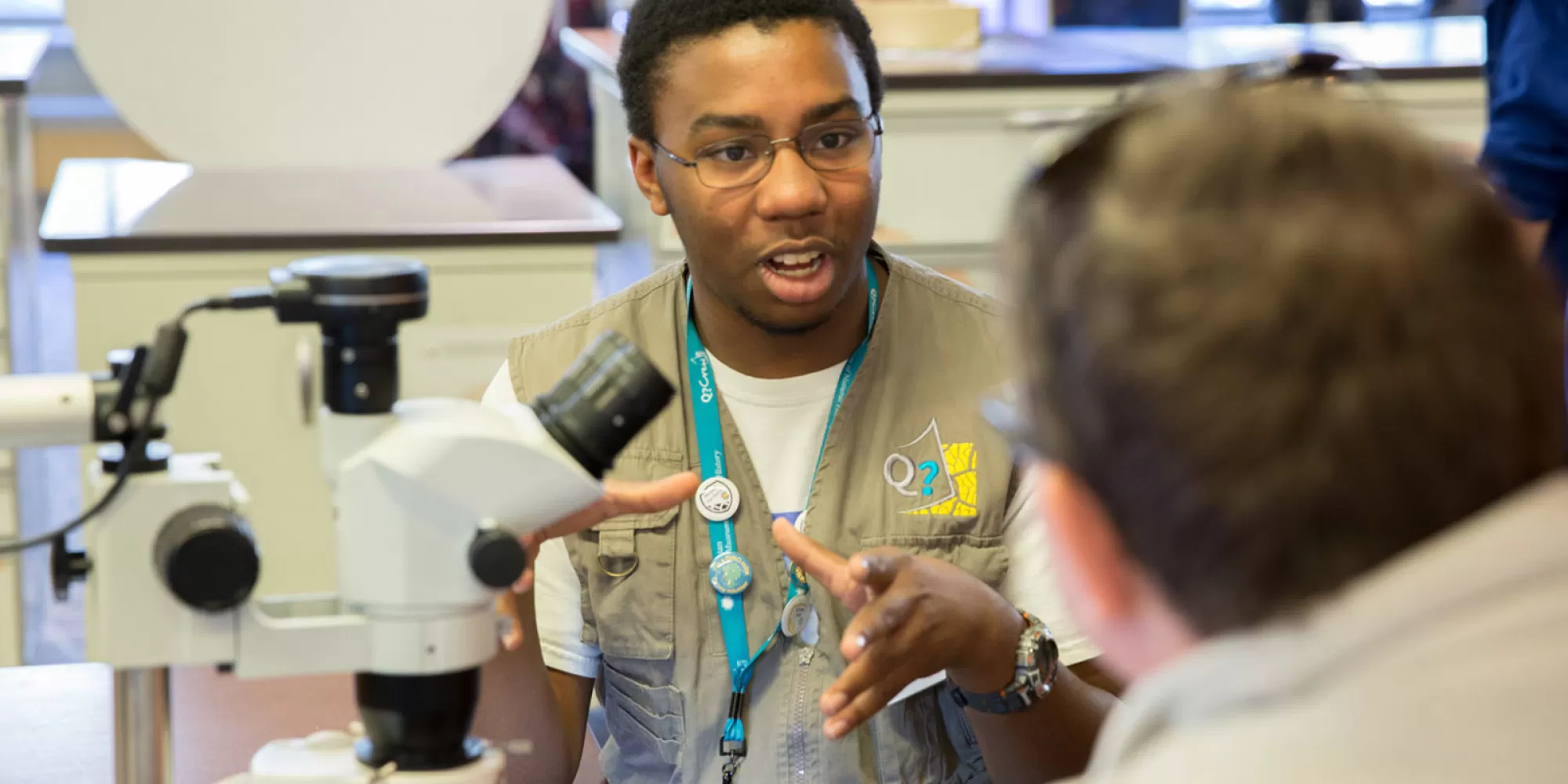 A Q?Crew teen volunteer sitting by a microscope, talking to a visitor.