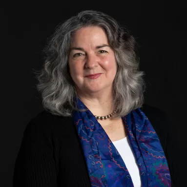 A headshot of Carol Butler, a white woman with gray hair wearing a white shirt, black sweater and colorful scarf. 