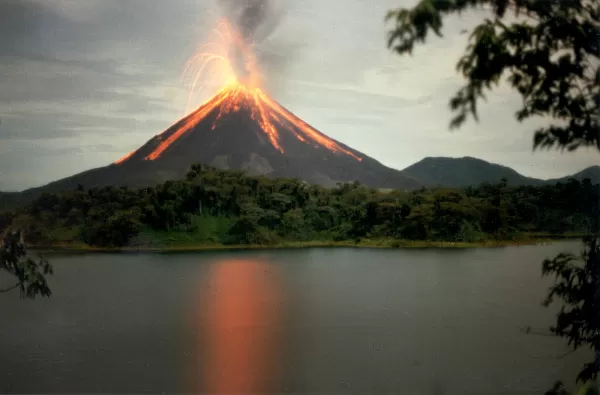 Volcano erupting with lava streaming down the side. A forest is at its foot and the eruption is reflected in a dark lake. 