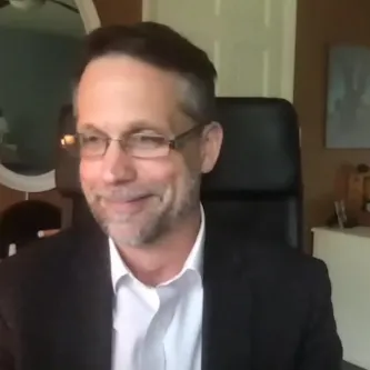 Frank Niepold, a light-skinned man wearing glasses and a black blazer, sitting in front of an oval mirror on the wall behind him