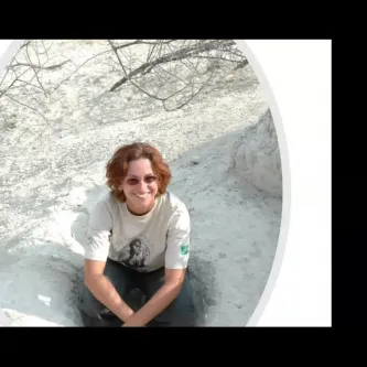 A light-skinned woman wearing sunglasses, a t-shirt, and hiking pants, squatting in a shallow hole in a dry, sandy white landscape.