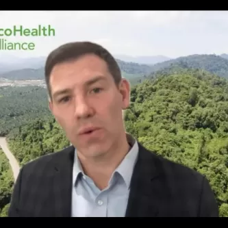 A man wearing a light-colored collared shirt and a dark sport coat. Behind him is a photo background of a forest.