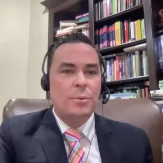 A man in a dark jacket, white shirt, and colorful striped tie sitting in a chair with a full bookcase behind him. He has a tan complexion, short dark hair, and is wearing a headphones with a headset microphone.