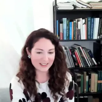 A light-skinned woman with long brown hair, sitting in front of a large bookcase full of books. She is wearing a white shirt with a pattern of large black and maroon flowers on it