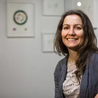 Ellie Irons in front of a wall with framed artwork on it. She is a white woman with long brown hair and is wearing a blue-gray sweater over a gray t-shirt