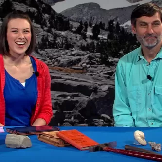 Maggy Benson and Dr. Brian Huber are seated at a table with several rocks of various sizes, a book, and several tools.