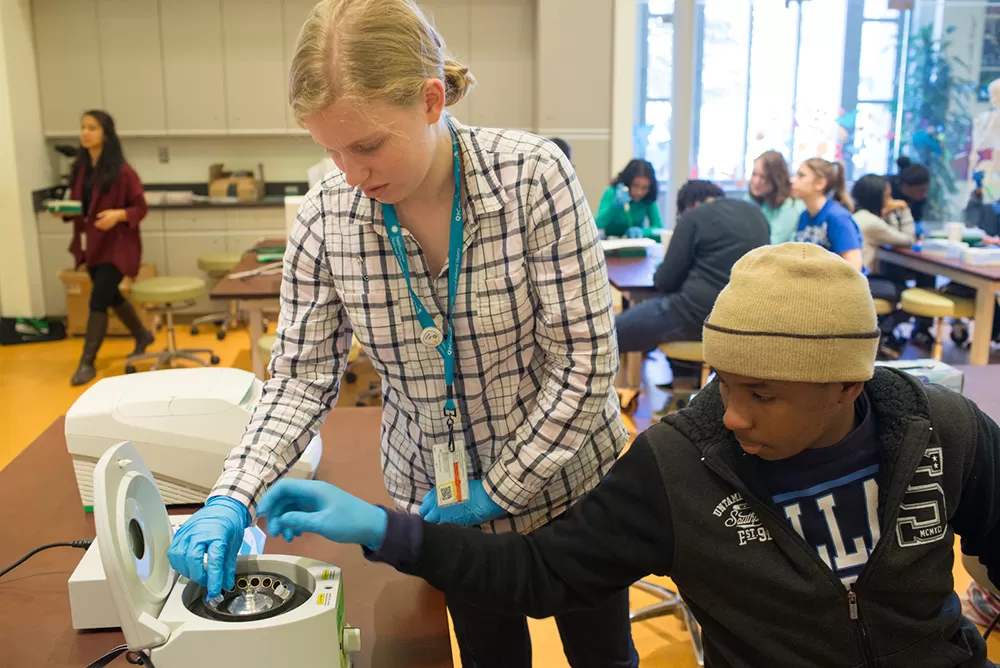 A teen girl and boy, wearing blue gloves, put tubes into a centrifugre.