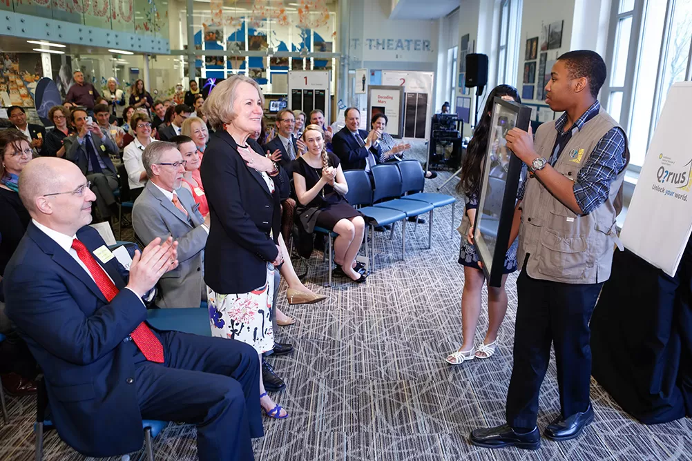 Corayln Whitney, a blonde woman in a blue blazer, stands with hand on heart as two teens present her with a framed picture.