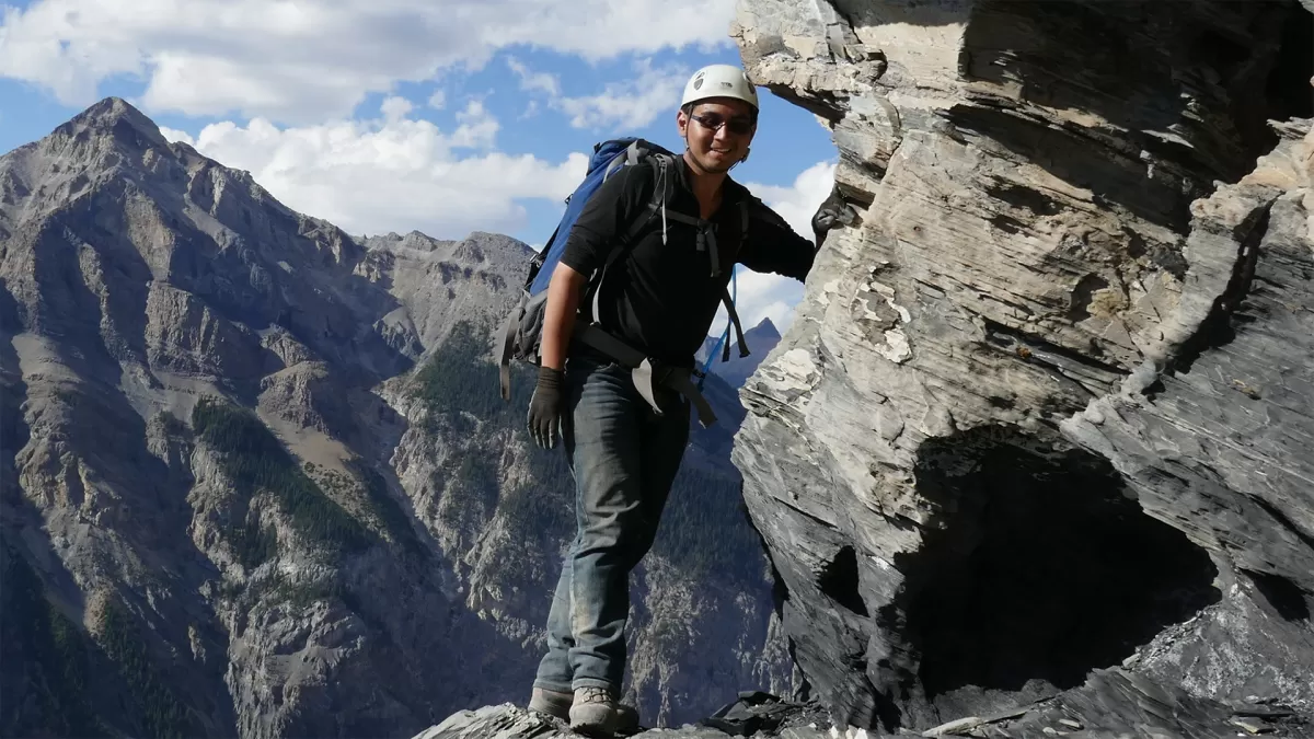 A man leaning against a rock face while wearing a helmet and backpack. Mountains are in the background.